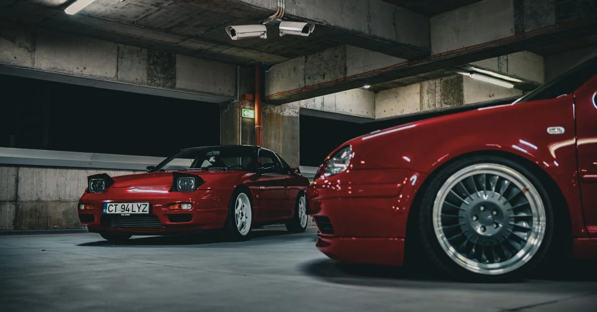 Capture de deux voitures vintage rouges garées dans un garage de béton urbain, éclairées par des lumières au plafond.