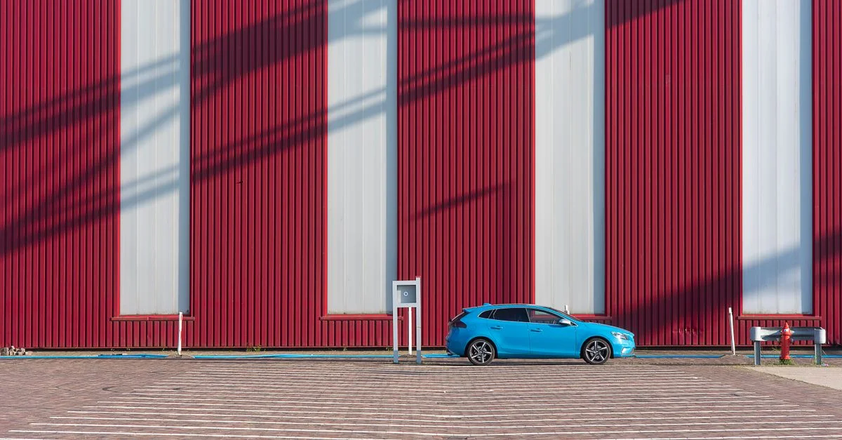 Une voiture d'un bleu éclatant garée contre un mur rouge vif sur un parking désert, mettant en valeur un design minimaliste.